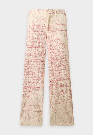 Homme portant une chemise crème avec un col rouge et des illustrations d'oiseaux, assortie à un pantalon large crème couvert de griffonnages rouges et de texte.