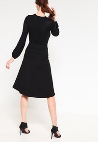 Haut noir à manches longues avec une texture côtelée et une jupe noire évasée, porté avec des chaussures à talons noires. La tenue est ajustée et élégante.