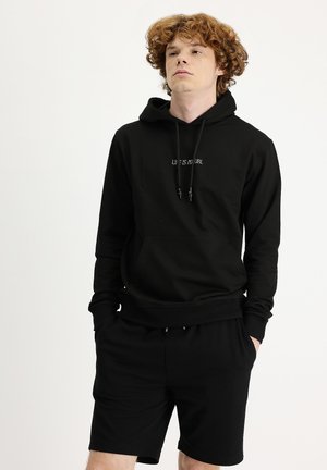 Sudadera negra con el texto "LA VIDA ES RURAL", bolsillo canguro y cordones ajustables. Combinada con pantalones cortos negros, ambos de una textura suave.