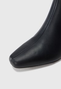 Μαύρο δερμάτινο μποτάκι με τετράγωνη μύτη και κεντρική ραφή, εμφανιζόμενο σε κοντινή λήψη πάνω σε γκρι ανοιχτό φόντο.