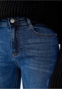 Jeans de mezclilla de color azul oscuro con corte ajustado, con costuras en contraste, cierre de botón y bolsillos frontales. El material tiene una textura suave.