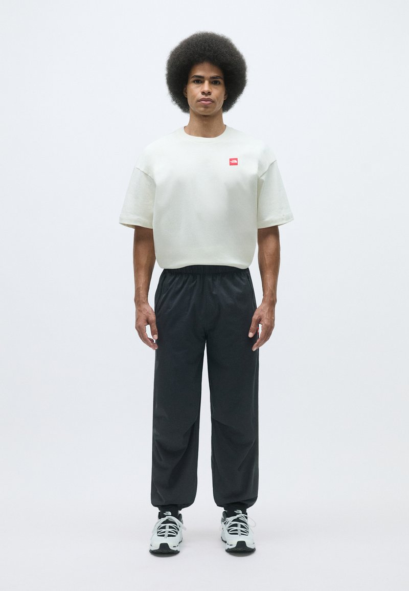 Ljusgrå oversized t-shirt med en liten röd logotyp, ihop med svarta byxor med elastisk midja och vita sneakers med svarta accenter.