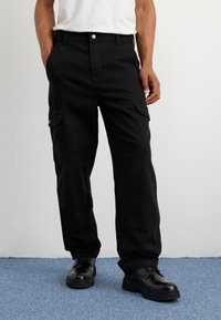 Μαύρο cargo παντελόνι με άνετη εφαρμογή, διαθέτοντας πλαϊνές τσέπες, βρόχεις ζώνης και κλείσιμο με κουμπί. Φοριέται με μαύρα κορδόνια παπούτσια.