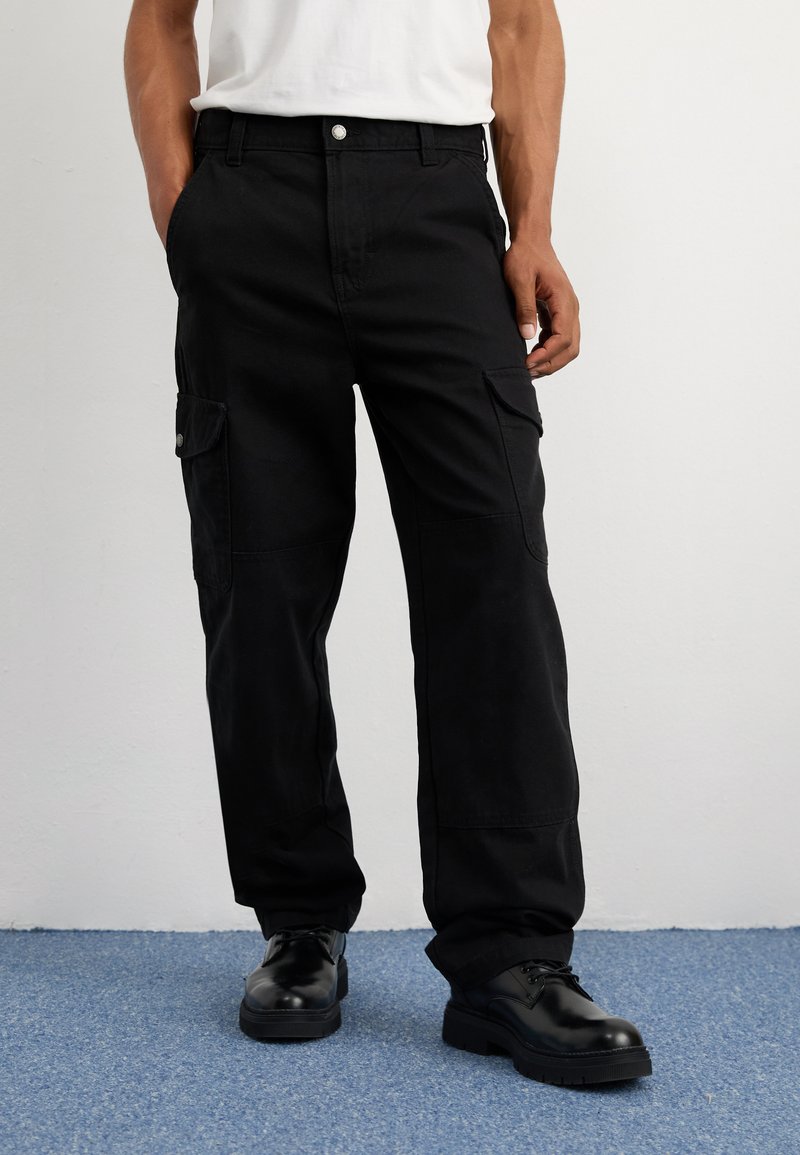 Μαύρο cargo παντελόνι με άνετη εφαρμογή, διαθέτοντας πλαϊνές τσέπες, βρόχεις ζώνης και κλείσιμο με κουμπί. Φοριέται με μαύρα κορδόνια παπούτσια.