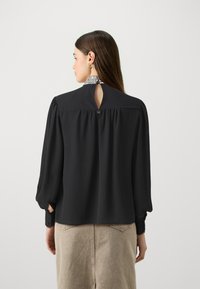 Blusa nera con maniche lunghe, apertura a chiave sul retro e dettagli plissettati. Presenta un colletto decorato e una texture liscia.