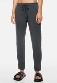 Grijze sweatpants met een elastische tailleband en een verstelbaar trekkoord. Gespen bij de enkels en een soepele textuur. Draagt met zwarte schuifslippers.