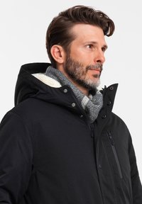 Giacca nera a isolamento termico con cappuccio foderato in pelliccia. Presenta una chiusura con zip e bottoni a pressione, tasche laterali e una sciarpa grigia a fantasia. Tessuto texture.