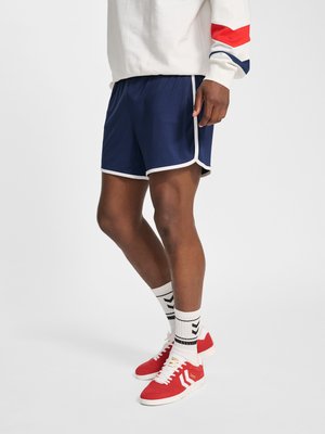 Mann trägt marineblaue Laufshorts mit weißem Besatz, weiße Socken mit schwarzen Chevron-Streifen, rote Turnschuhe und einen weißen Pullover mit roten und blauen Armbändern.
