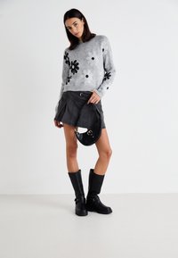 Grau gestrickter Pullover mit schwarzen Blumenmustern, kurzer schwarzer plissierter Rock, schwarze kniehohe Stiefel und eine kleine schwarze strukturierte Tasche.