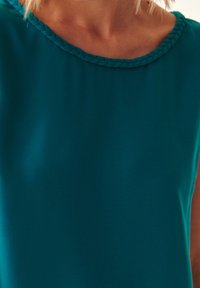Top sin mangas en color verde azulado con escote redondeado, que presenta un detalle trenzado en el borde. Textura suave con un ajuste relajado.