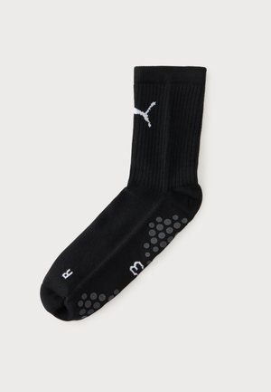 Chaussette de sport noire avec un logo Puma blanc, des picots antidérapants gris sur la semelle et les lettres blanches “L” et “M” près des orteils, sur un fond uni.