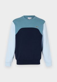 Sweat-shirt col rond avec un design en blocs de couleurs comprenant des matériaux bleu marine, bleu clair et bleu sarcelle. Poignets et ourlet côtelés. Logo Lacoste sur la poitrine.