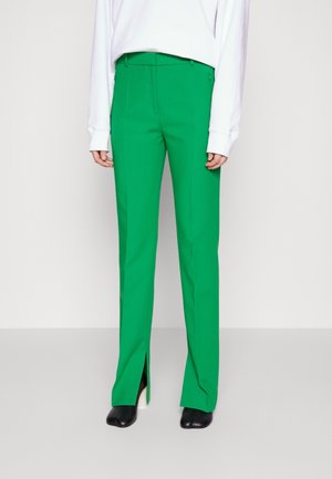 Personne portant un pantalon vert vif sur mesure avec des fentes à l'avant, associé à un haut blanc à manches longues et des chaussures à talons noires sur un fond uni.