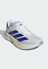 Zapatilla deportiva blanca con acentos morados, parte superior de malla texturizada, cuello acolchado y suela ligera con detalles moldeados.