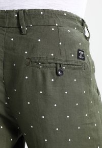 Bavlněné kalhoty olivově zelené barvy s bílými puntíky, s knoflíkovým zapínáním zadní kapsy a tkanou štítkem. Textura působí lehce a vzdušně.