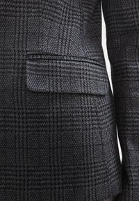 Poche à rabat sur le côté droit d'un blazer à motif pied-de-poule gris foncé et noir.