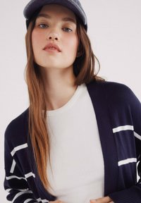 Námořnický kardigan s bílými pruhy přes bílou žebrovanou košili. Měkká tkanina, otevřený design vpředu a jednoduchý kulatý výstřih.