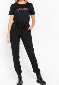 Vrouw in een zwarte t-shirt met korte mouwen met de tekst "Happy", hoge zwarte broek met een strikceintuur en zwarte enkellaarsjes, poseert met een hand in de zak.