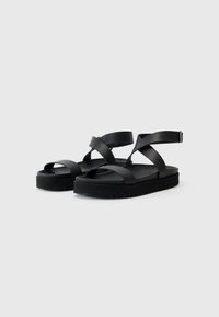 Sorte svarte sandaler med kryssremmer laget av glatt syntetisk materiale, med en polstret fotseng og teksturert gummisåle.