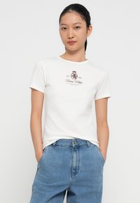 Tricou alb cu mânecă scurtă, având un logo imprimat de culoare maro închis. Materialul pare moale și are o croială clasică. Este asortat cu blugi din denim albastru.