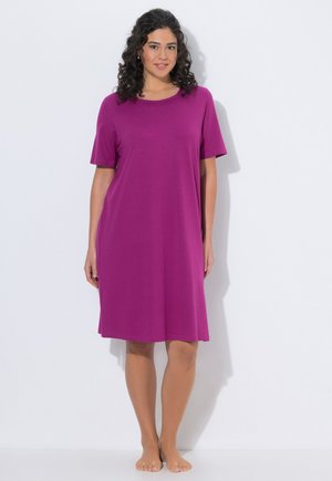 Kvinde med krøllet hår stående barfodet i en løs, knælængde, kortærmet lilla kjole mod en ensartet lys baggrund.