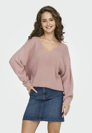 Maglione lavorato a maglia rosa chiaro con scollatura profonda a V e maniche lunghe larghe, abbinato a una gonna in denim blu. La texture è spessa e rilassata.
