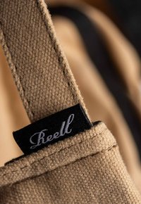 Correa de tela beige con un tejido texturizado y un borde cosido, resaltando una etiqueta negra con texto en estilo manuscrito blanco.