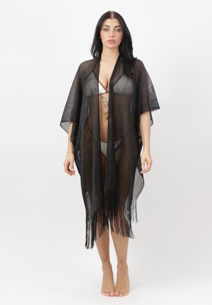 Kvinde iført en gennemsigtig sort frynset overdel over en hvid bikini, stående barfodet mod en ensfarvet hvid baggrund.