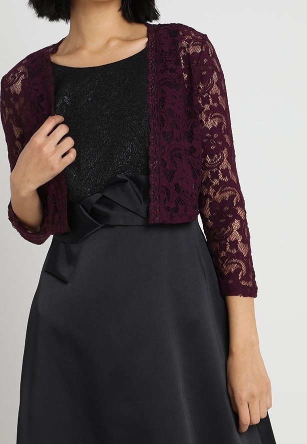 Femme portant une robe en satin noire avec un corsage texturé et un cardigan court en dentelle violet foncé à manches trois-quarts.