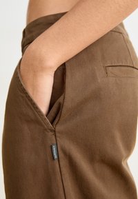 Pantalon en coton marron avec une poche latérale, présentant des coutures visibles et une petite étiquette de marque sur le bord de la poche. Texture lisse.