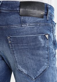 Denim jeans in dark blue with a faded finish. Features a black leather label on the waistband and decorative metal accents on pockets.