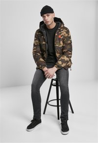 Kurtka puchowa w kamuflażu z czarnym wnętrzem, noszona na czarnym t-shircie, w połączeniu z ciemnymi dżinsami i czarnymi sneakersami. Czarna czapka na głowie.