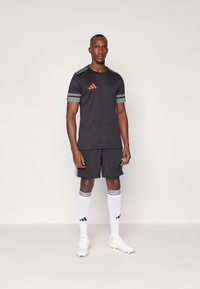 Schwarzes Athletik-Trikot mit rotem Logo und grauen Streifen, kombiniert mit schwarzen Shorts, weißen Socken mit schwarzen Akzenten und weißen Fußballschuhen.