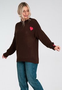 Brązowy sweter dziany o luźnym fasonie, z różowym sercem na piersi. Długie rękawy i ściągacze wykańczające dopełniają całość projektu.