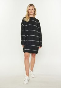Femeie stând în picioare, purtând o rochie tricotată neagră cu dungi albe și adidași albi, pe un fundal simplu deschis.