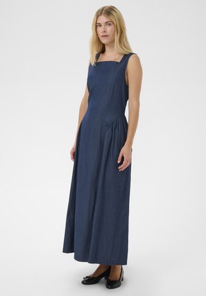 Szőke nő ujjatlan, négyzet nyakú sötétkék maxi ruhában és fekete alacsony sarkú cipőben, lazán tartja karjait, fehér háttér előtt áll.