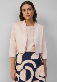 Lichtroze cropped jasje met driekwartmouwen, over een bleke top en een marineblauwe rok met grote geometrische patronen in crème en rood.