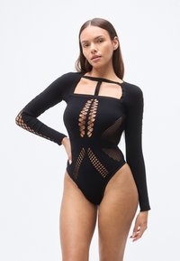 Μαύρο κορμάκι με μακριά μανίκια και ανοίγματα, με τετράγωνη λαιμόκοψη και λεπτομέρειες από δίχτυ αλιείας κατά μήκος των χεριών και του κορμού. Λείο ύφασμα.