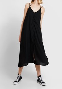 Robe noire sans manches avec un décolleté en V, fermeture à boutons sur le devant et fentes sur les côtés, associée à des baskets montantes noires.