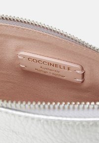 Hopeapintainen nahkapussukka, jossa näkyvä vetoketju, pinkki kangaspinta ja tagi, jossa lukee "COCCINELLE Aito italialainen nahka."
