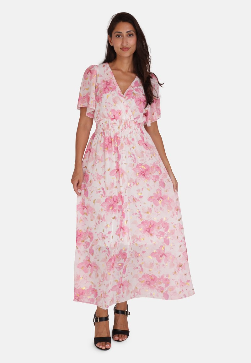 Robe maxi avec des manches courtes flottantes, motif floral rose sur un fond clair, cintrée à la taille, en tissu transparent, portée avec des talons noirs.
