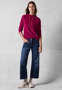 Felpa fucsia con cappuccio e lacci, abbinata a jeans dritti scuri a vita alta. Sneakers color tan completano l'outfit.