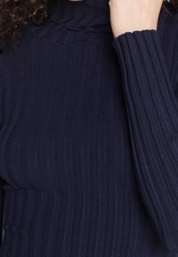 Pull à col roulé côtelé bleu marine à manches longues porté par une personne aux cheveux bouclés, la main posée près du cou.