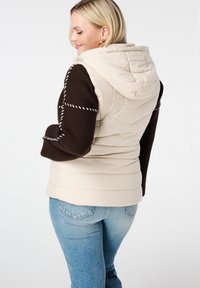Beige puffer vest met een capuchon, met donkerbruine gebreide mouwen en witte stikselaccenten. De pasvorm is relaxed, met een gestructureerd ontwerp.