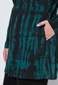 Zwarte tuniek met lange mouwen en een teal abstract patroon, met een zijzak en een relaxte pasvorm. Gemaakt van zachte, rekbare stof.