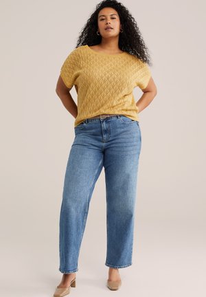 Kvinde med krøllede hår står med hænderne bag ryggen iført en gul tekstureret kortærmet top, blå jeans og beige hæle.