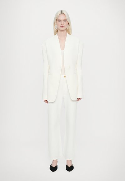 Costume blanc ajusté avec un blazer structuré, des manches longues et un pantalon à jambes larges. Texture lisse ; fermeture à un bouton. Chaussures noires à bout pointu.