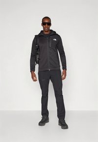 Μαύρο ζακέτα με φερμουάρ και αντίστοιχο παντελόνι από ελαφρύ ύφασμα. Το μοντέλο φοράει σκούρα γυαλιά ηλίου και μαύρα ορειβατικά μποτάκια. Φορητό σακίδιο πλάτης προσαρτημένο.