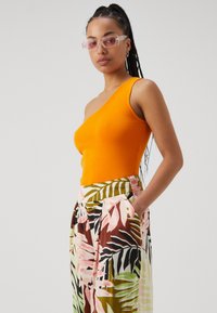 Top naranja de canalé con un hombro, combinado con pantalones de pierna ancha de estampado floral multicolor que cuentan con bolsillos y una mezcla de tonos rosa, verde y marrón.