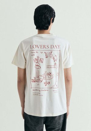 Persona que lleva una camiseta blanca con un diseño en línea roja de una mesa, flores, un querubín y el texto "DÍA DE LOS AMANTES" en la espalda.